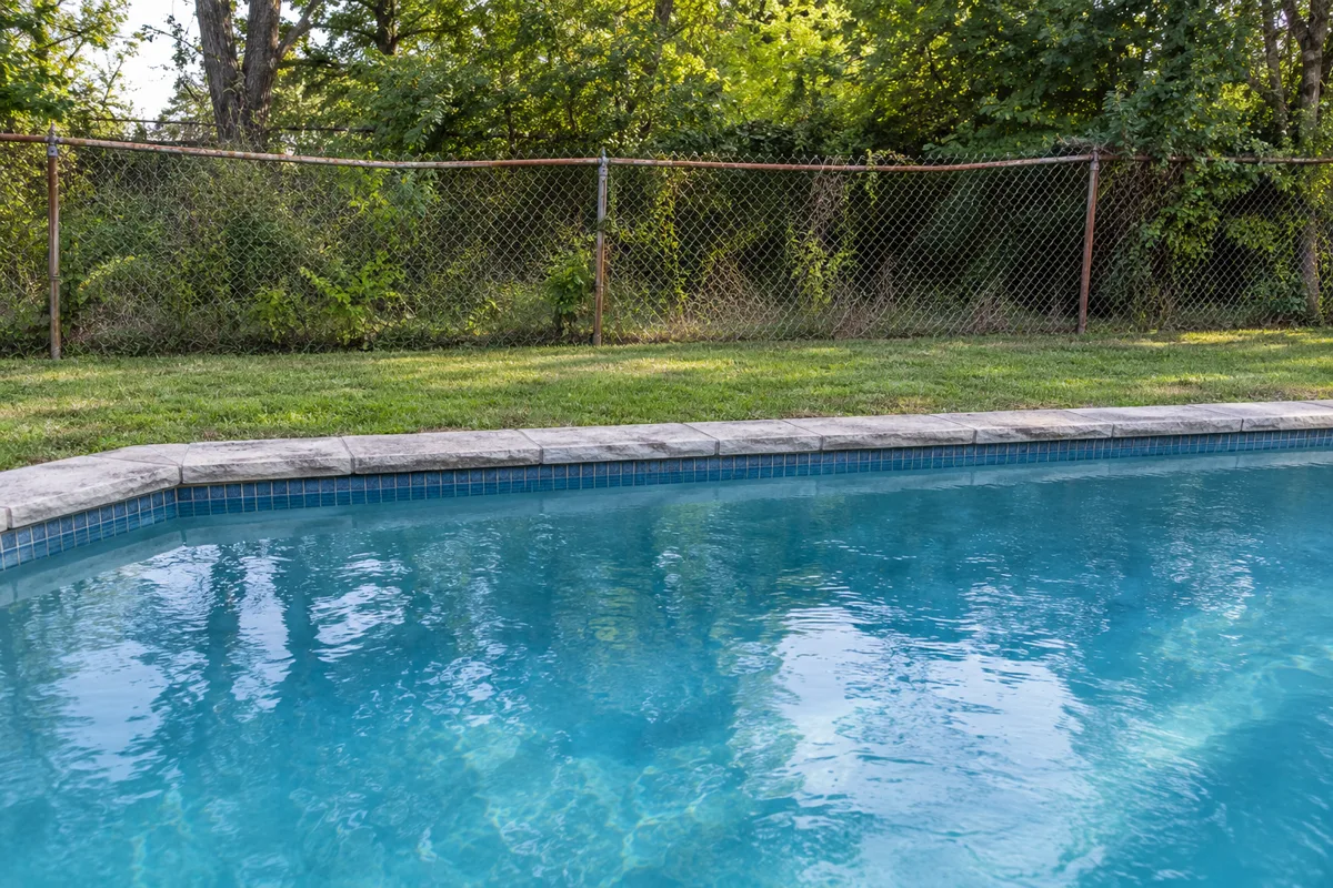 Pool edge with stone coping and a lawn, backed by a rusted, sagging chain-link fence in front of trees